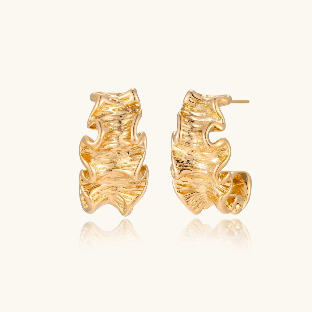 Pair of gold huggie hoops with sculpted wave texture and dimensional ripples on neutral background.