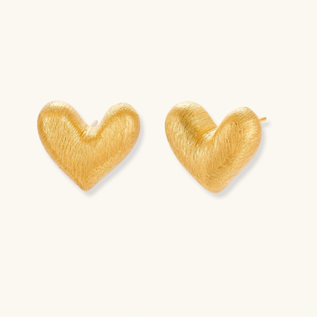 Pair of oversized brushed gold heart stud earrings with a puffy, sculpted surface and post-back closure on a cream background.