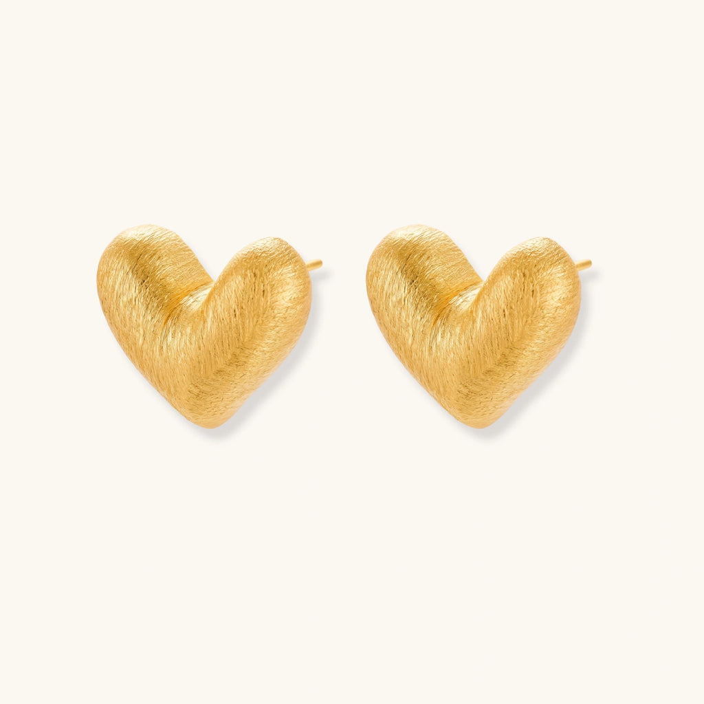 Pair of oversized brushed gold heart stud earrings with a puffy, sculpted surface and post-back closure on a cream background.