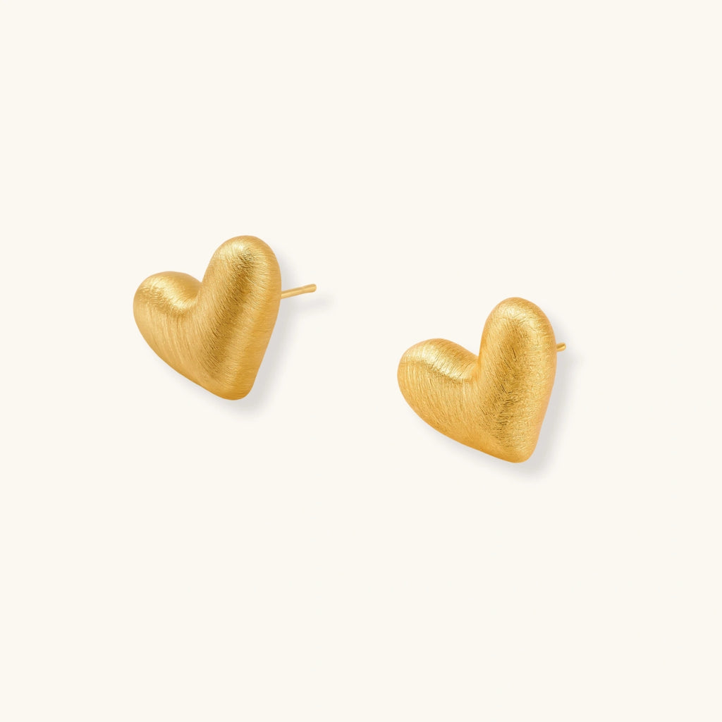 Pair of oversized brushed gold heart stud earrings with a puffy, sculpted surface and post-back closure on a cream background.