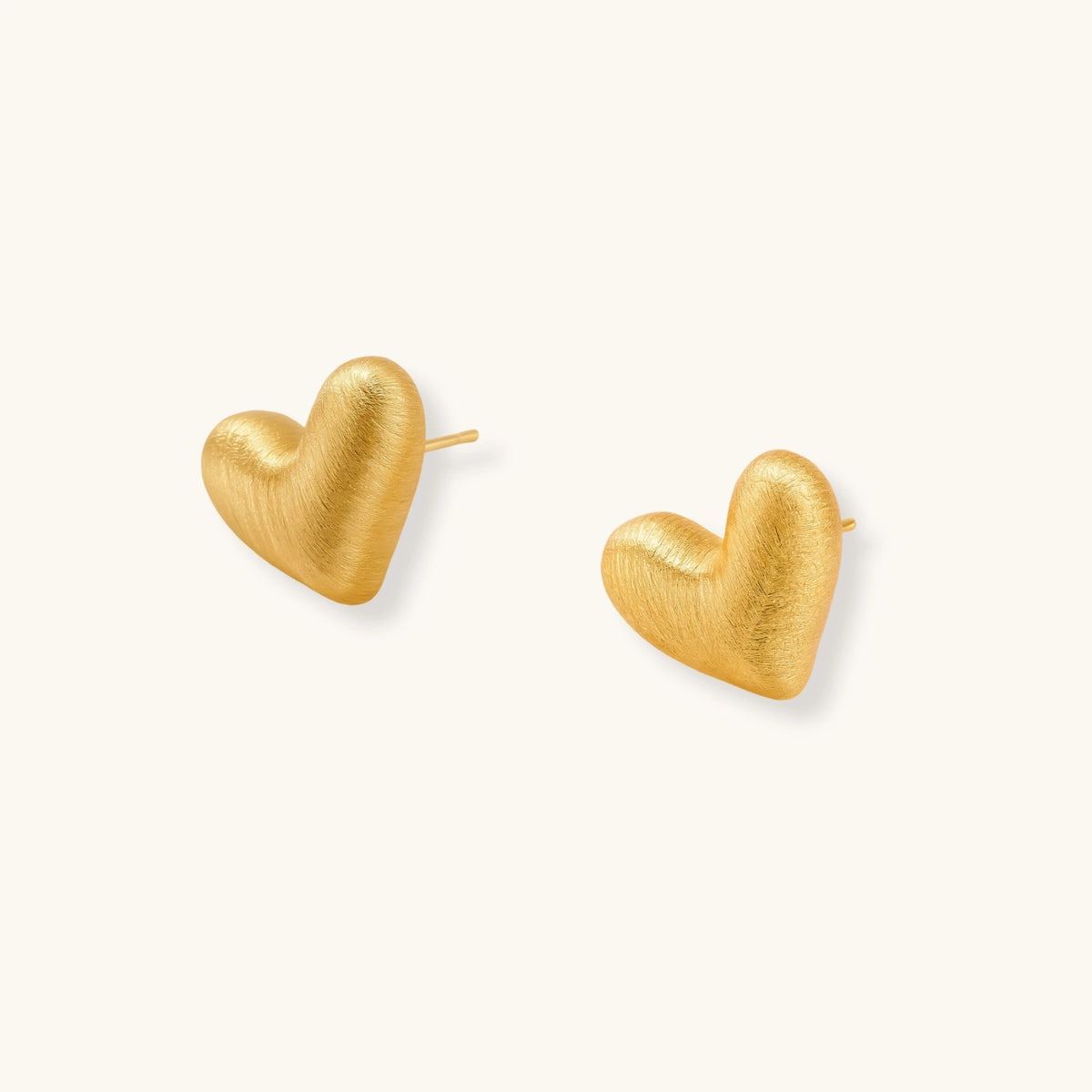 Pair of oversized brushed gold heart stud earrings with a puffy, sculpted surface and post-back closure on a cream background.